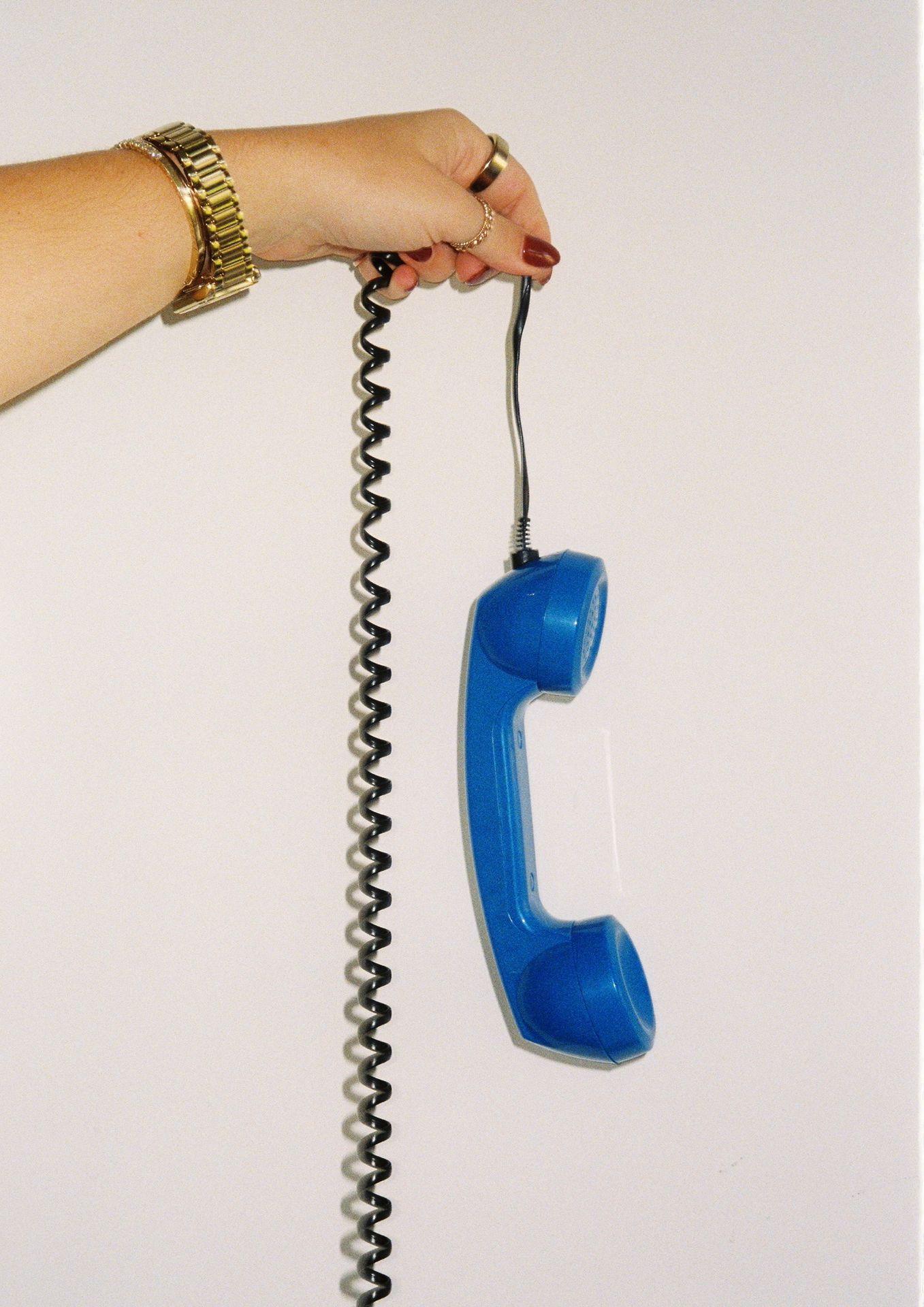 Hand holding a blue retro phone receiver with coiled cord against a plain background.